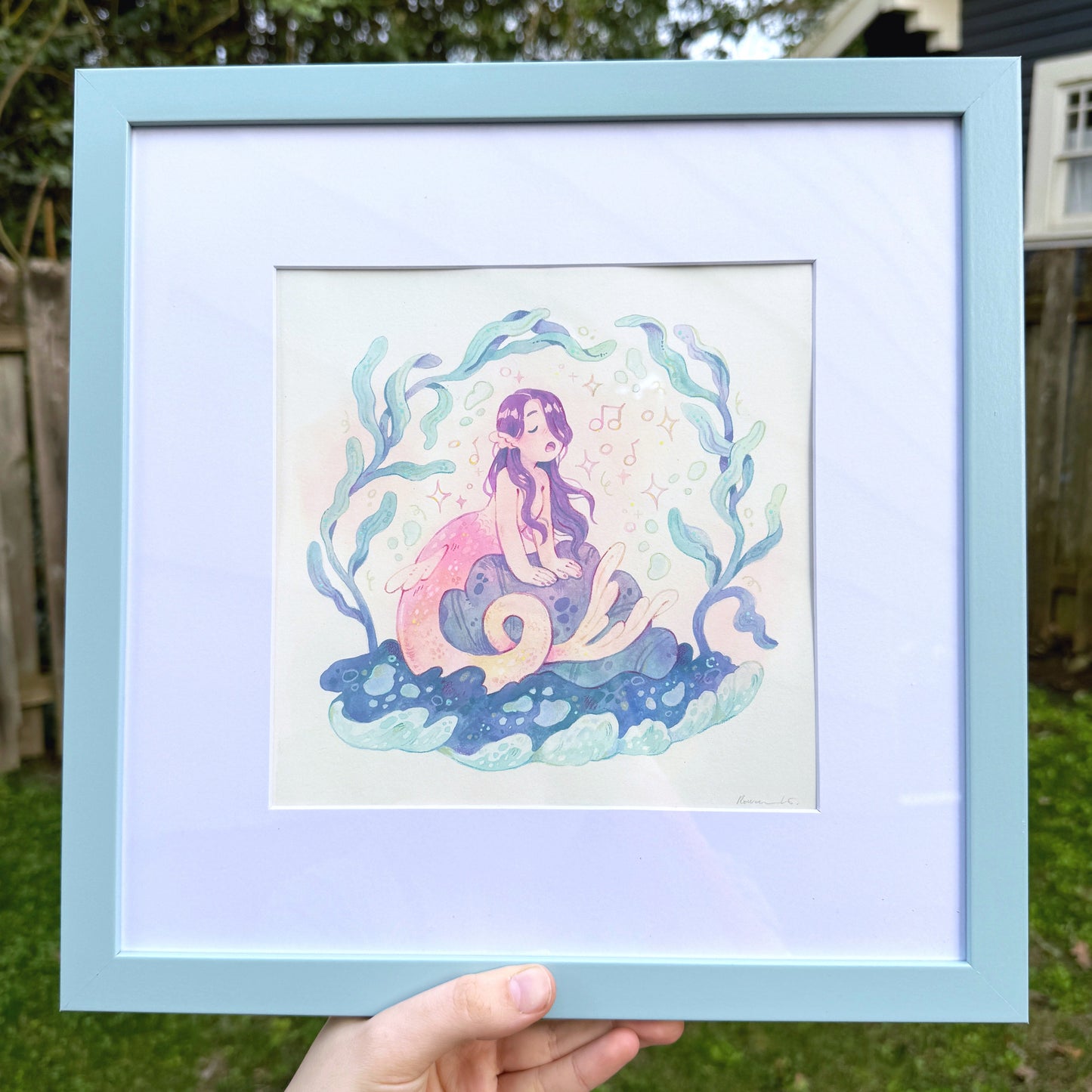 Framed artwork of a mermaid held in front of a natural background